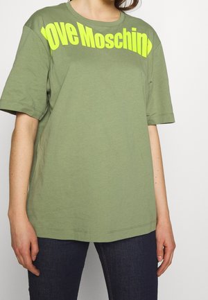Personne portant un T-shirt oversize vert olive avec un texte jaune néon « Love Moschino » sur la poitrine et un jean bleu foncé.