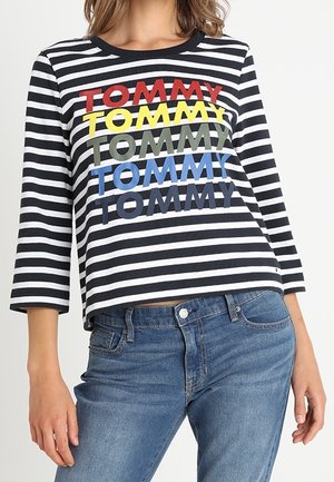 Een zwart-wit gestreept shirt met lange mouwen, bedrukt met "TOMMY" vijf keer in rood, geel, groen, blauw en marineblauw, gedragen met een blauwe spijkerbroek.