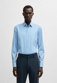 Camicia azzurro chiaro a maniche lunghe con bottoni, caratterizzata da righe verticali e colletto a punta, abbinata a pantaloni scuri con texture, dal taglio sartoriale.