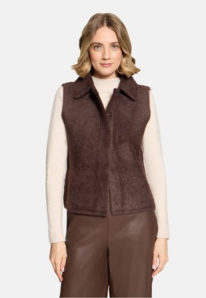 Femme portant un gilet sans manches marron foncé duveteux sur un haut crème à manches longues et un pantalon en cuir marron, debout devant un fond blanc.