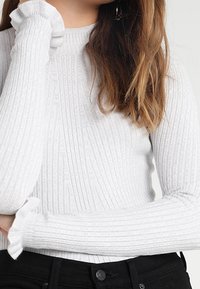 Vaaleanharmaa ribbineulottu paita, jossa pitkä hihat. Siinä on korkea pääntie ja teksturoitu kuvio. Käytetty myös kerätty hiusresoriyksityiskohta.