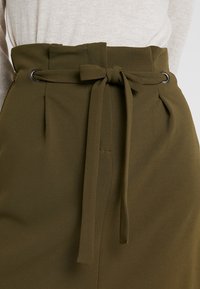 Jupe taille haute vert olive avec ceinture, réalisée en tissu texturé. Dispose de plis et de passants métalliques pour la fermeture.