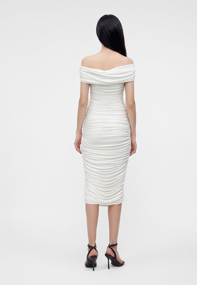 Kvinde med langt sort hår iført en hvid off-shoulder midi kjole med rynker og sorte høje hæle sandaler, stående med ryggen mod.