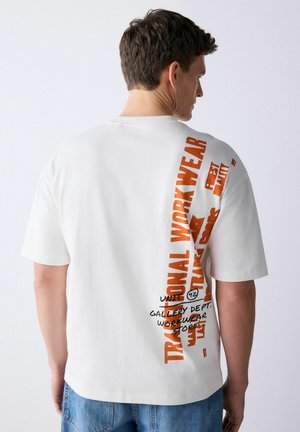 Bărbat purtând tricou alb cu text vertical portocaliu „Traditional Workwear” și text negru în stil de scris de mână pe spate, asortat cu blugi albaștri.