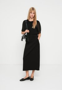 Schwarzes Kleid mit kurzen Puffärmeln, tailliertem Bund und geradem Maxilänge. Kombiniert mit schwarzen Ballerinas und einer kleinen schwarzen Handtasche.