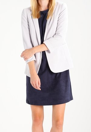 Femme portant une robe bleu marine et un blazer gris clair avec les manches retroussées, debout, les bras croisés devant un fond blanc.