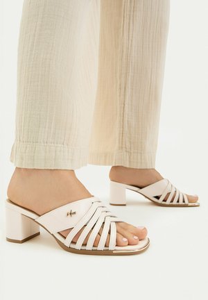 Fødder iført beige sandaler med blokhæle og stropper, kombineret med lette, teksturerede, vidde bukser på en neutral baggrund.