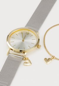 Montre en argent ornée d’accents dorés avec bracelet en maille et pendentif cœur, à côté d’un bracelet doré assorti avec pendentif cœur sur fond blanc.