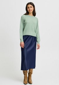 Hellgrüner Strickpullover mit langen Ärmeln, kombiniert mit einem dunkelblauen Satin-Midirock und hellbraunen kniehohen Stiefeln. Schlichtes Design und glatte Textur.