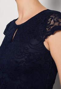 Gros plan d'une femme portant une robe en dentelle bleu marine avec des manches courtes et un petit décolleté en forme de trou de serrure.