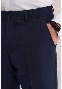 Moški v mornarsko modrih elegantnih hlačah s stranskimi žepi, beli srajci zatlačeni noter in črni usnjeni ročni uri na zapestju.