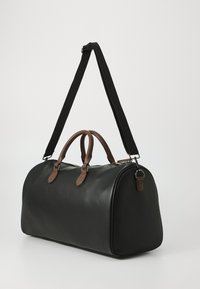 Bolsa de viaje negra con acentos en piel marrón, forma redondeada, asa superior de tela y correa para el hombro negra desmontable. Textura suave.