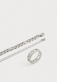 Conjunto de joias de metal prateado com uma pulseira de corrente grossa, uma corrente delicada e um anel texturizado com aberturas, todos com um acabamento polido.