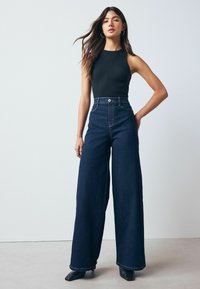 Černý žebrovaný top na ramínka spárovaný s vysokými, širokými džínami z temného denimu. Modelka nosí špičaté černé lodičky. Jednoduchý, čistý design.