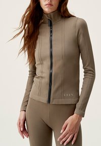 Giacca sportiva marrone con zip e colletto alto, motivo testurizzato e zip nera. Leggings marroni abbinati visibili sotto.
