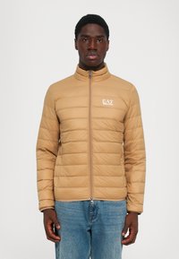 Beige steppelt dzsekit viselő férfi, elöl cipzárral és EA7 Emporio Armani logóval, kék farmerrel párosítva, egy sima háttér előtt néz előre.