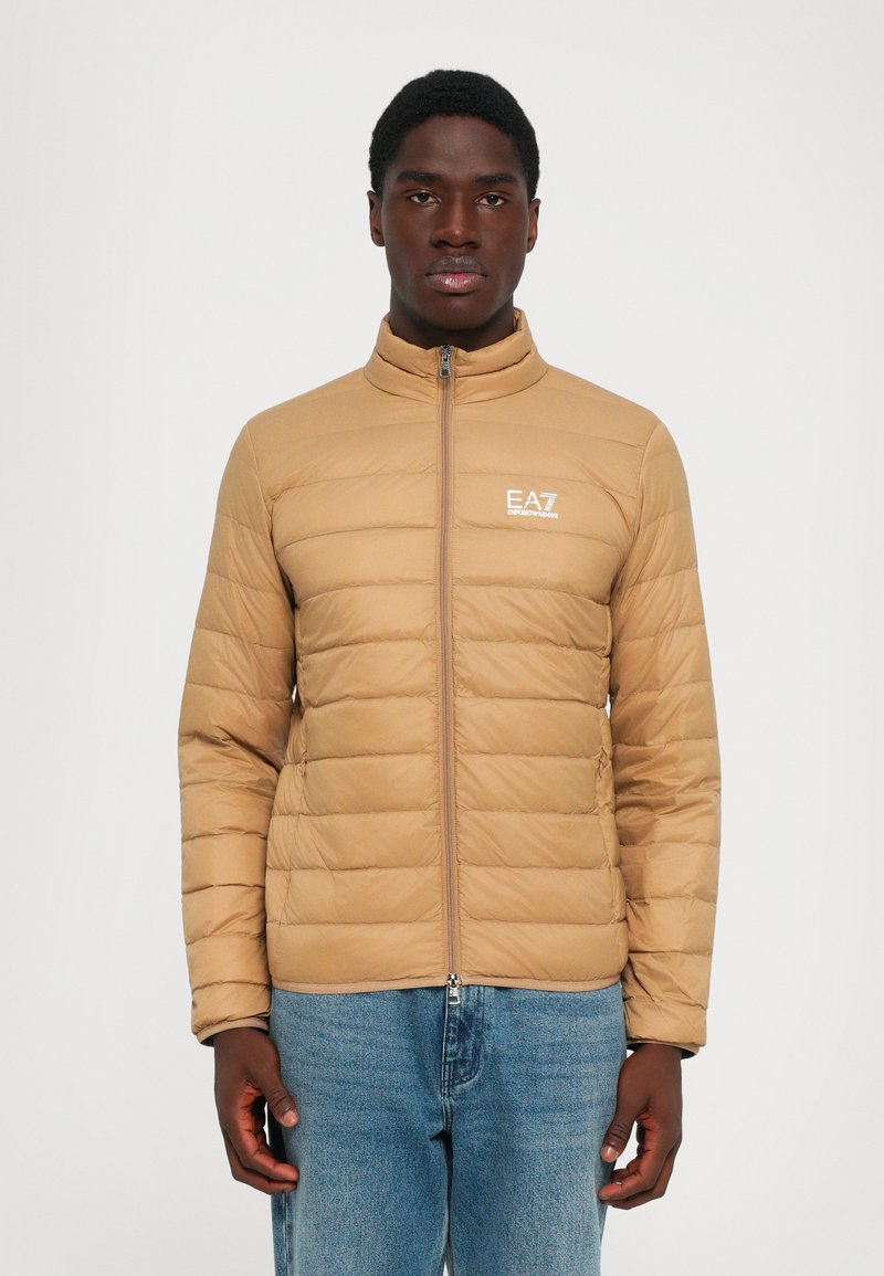 Beige steppelt dzsekit viselő férfi, elöl cipzárral és EA7 Emporio Armani logóval, kék farmerrel párosítva, egy sima háttér előtt néz előre.