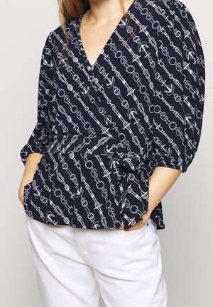 Vrouw draagt een marineblauwe wikkelblouse met wit nautisch touw, ankerpatroon en ballonmouwen, gecombineerd met witte broek, handen in de zakken.