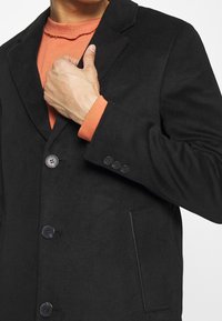 Černý blazer z vlněné směsi s klopami, třemi předními knoflíky a jednou náprsní kapsou. Vnitřní vrstva obsahuje detail z oranžové látky.