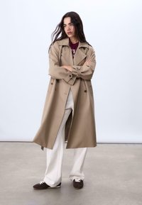 Cappotto trench beige doppiopetto con risvolti larghi, maniche lunghe e tasche frontali, indossato sopra una camicia bordeaux e pantaloni bianchi.