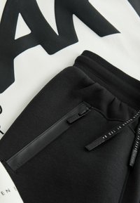 Fekete melegítőnadrág cipzáras zsebbel, struktúrált anyaggal és márkajelzésekkel. A háttérben egy fehér ing látható vastag fekete felirattal.