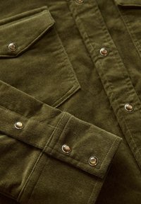 Olijfgroene corduroy shirt met twee borstzakken, zilveren drukknoopjes en een gestructureerde stof. Close-upweergave benadrukt de stikdetails.