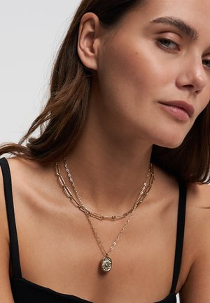Woman wearing two layered gold chain necklaces, one with a rectangular linked chain and the other with a sun pendant, dressed in a black top.