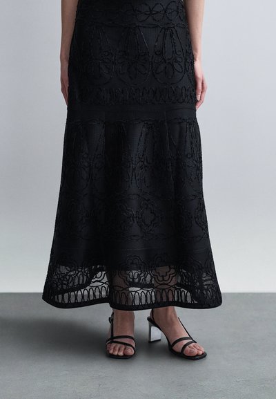Femme portant une jupe midi en dentelle noire à motifs floraux et des sandales noires à talons blocs avec lanières, debout sur un sol gris contre un mur uni.