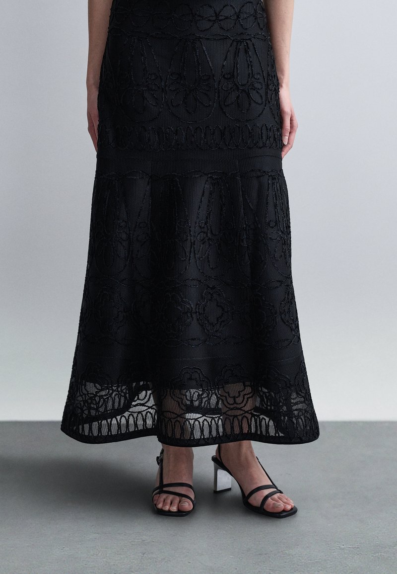 Femme portant une jupe midi en dentelle noire à motifs floraux et des sandales noires à talons blocs avec lanières, debout sur un sol gris contre un mur uni.