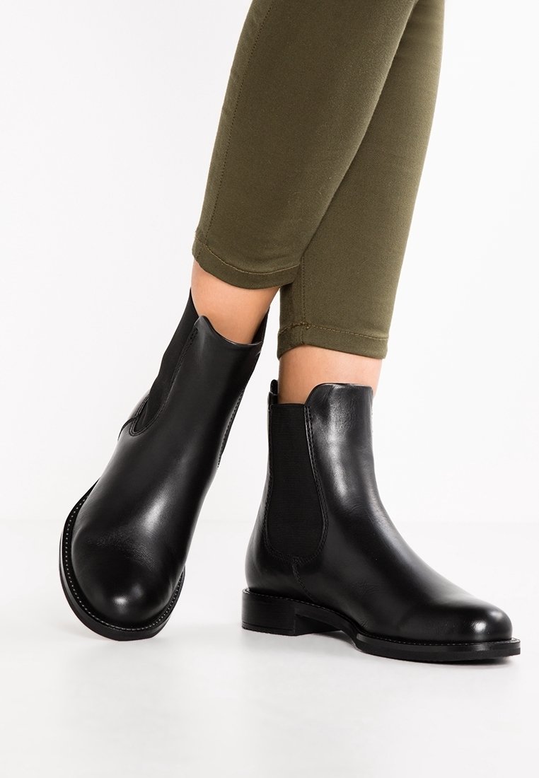Bottines en cuir noir avec panneaux latéraux élastiques portées avec un pantalon ajusté vert olive sur fond blanc.