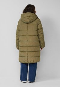 Olivgrüner Puffer-Mantel mit Kapuze, ausgestattet mit gesteppten Abschnitten und einem mittellangen Schnitt. Kombiniert mit blauen weiten Jeans und weißen Sneakers.