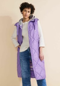 Lila kviltad lång väst med grå huva, tryckknappar och sidofickor, bärs över en krämfärgad tröja och blå jeans. Slätt, lätt material.