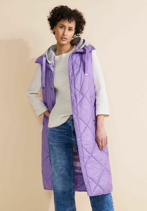 Lila gesteppte lange Weste mit grauem Kapuzen, Druckknöpfen und seitlichen Schlitzen, getragen über einem cremefarbenen Pullover und blauen Jeans. Glattes, leichtes Material.