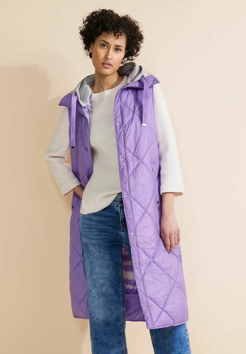 Lila kviltad lång väst med grå huva, tryckknappar och sidofickor, bärs över en krämfärgad tröja och blå jeans. Slätt, lätt material.