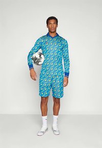 Ung mann iført blå og grønn fotballdrakt med geometrisk mønster, holder en svart-hvitt fotball under armen og står mot en ensfarget bakgrunn.