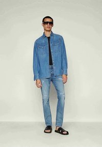 Moški z temnimi sončnimi očali, modro denim srajco, črno majico, raztrganimi svetlo modrimi kavbojkami in črnimi sandali brez prstov stoji ob enobarvni ozadju.