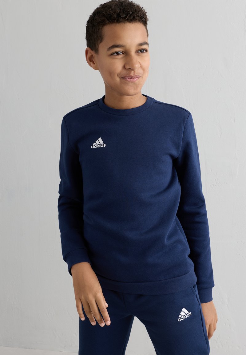 Granatowa bluza typu sweatshirt z okrągłym dekoltem i długimi rękawami, z białym logo Adidas na lewej piersi. Wykonana z miękkiego materiału o gładkiej fakturze.