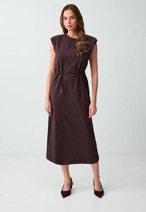 Donna con lunghi capelli mossi che indossa un vestito midi marrone scuro senza maniche con una sottile cintura e tacchi a punta, in piedi davanti a uno sfondo semplice.