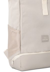 Gros plan sur un sac à dos beige clair, montrant une poche latérale, une poche zippée en bas et un écusson du logo Johnny Urban sur le devant.
