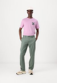 Lyserød kortærmet t-shirt med en sort og hvid grafisk lomme, parret med lysegrønne bukser og beige sportssko.