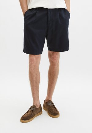 Mężczyzna ubrany w ciemnogranatowe szorty, białą koszulę i brązowe zamszowe buty z jasnobrązowymi podeszwami, stojący z rękami w kieszeniach.