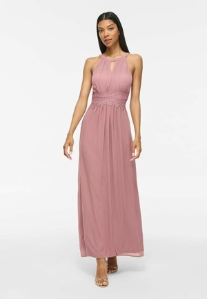 Femme portant une robe longue rose poudré sans manches avec un détail en dentelle à la taille et des talons dorés à brides, debout devant un fond blanc.