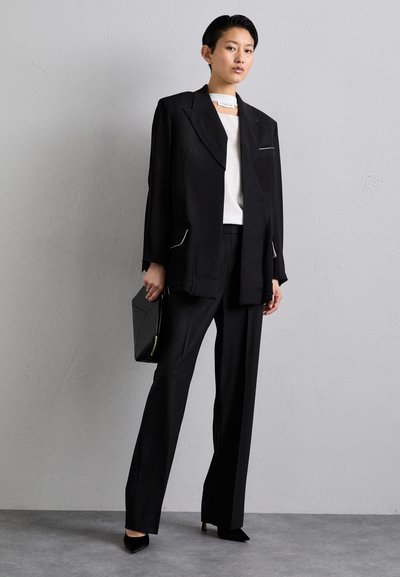 Black tailored suit with a loose-fit blazer featuring white accents, paired with a white top and straight-leg trousers, completed with pointed heels.