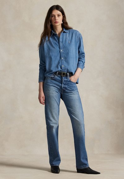 Vrouw met lang bruin haar die een blauw denim overhemd draagt, ingestopt in een hoge taille blauwe spijkerbroek en zwarte puntige schoenen, staand met één hand in de zak.