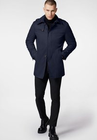 Granatowy płaszcz z wykładanym kołnierzem, z zapięciem na guziki i dwoma bocznymi kieszeniami; noszony na czarnym golfie i czarnych spodniach.