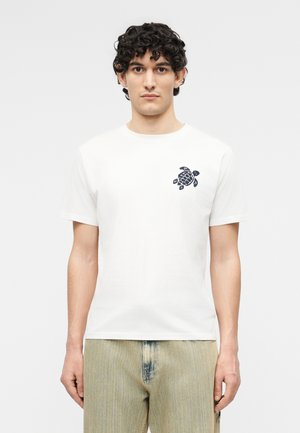 Young man with curly hair wears a white t-shirt featuring a small black sea turtle graphic on the left chest and light beige pants.