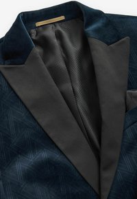 Marineblaue Jacke mit gemusterter Textur, Samtkragen und kontrastierendem schwarzen Revers. Das Innenfutter besteht aus Netzmaterial und es gibt ein Etikett am Kragen.