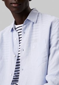 Camicia di lino azzurro chiaro con chiusura a bottoni, taschino sul petto e trama sottile. Indossata sopra una t-shirt bianca con righe blu navy.