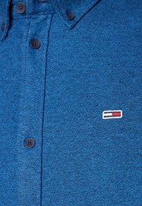 Blaues strukturiertes Hemd mit Button-Down-Kragen, ausgestattet mit schwarzen Knöpfen und einem kleinen gestickten Logo in rot, weiß und blau auf der Brust.