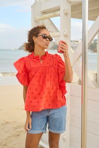 Femme en blouse rouge et short en jean tenant une canette de soda près d'une tour de sauveteur sur une plage ensoleillée avec une mer calme en arrière-plan.